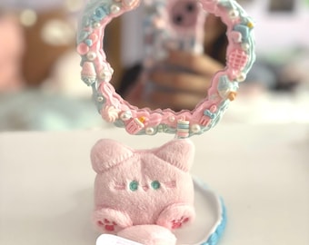 Pode incluir: Um espelho circular com uma moldura decorada em tons pastel de azul e rosa com textura de glacê, apresentando berloques em miniatura e pérolas brancas. A base é uma figura de gato de pelúcia rosa com detalhes bordados e um pequeno curativo.