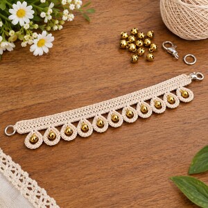 May include: A beige crocheted trim featuring a series of teardrop-shaped loops along the bottom edge, each containing a small, shiny gold-colored bell. The trim includes silver-toned metal lobster clasps and rings at both ends for attachment.