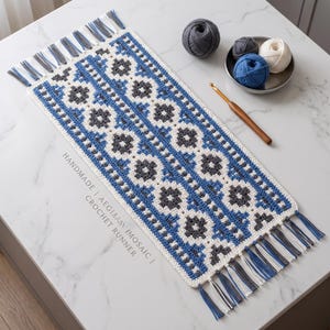 May include: A rectangular crochet runner featuring a geometric mosaic pattern in blue, white, and dark gray. The runner has fringe tassels on both ends in matching colors. Text on the runner reads HANDMADE | AEGEAN MOSAIC | CROCHET RUNNER.