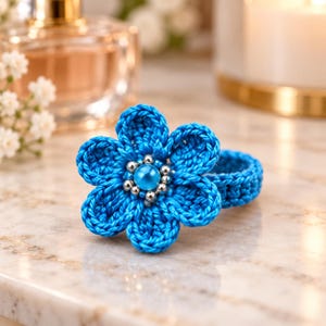 Puede incluir: Un accesorio para el cabello con forma de flor tejida a ganchillo en color azul brillante, con una cuenta azul claro central rodeada de pequeñas cuentas metálicas plateadas. La flor está unida a una banda tejida a juego, creando una forma circular.