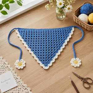 Puede incluir: Un pañuelo triangular de ganchillo en tono azul intenso con un patrón de puntos calados y un borde festoneado blanco. Los dos lazos largos terminan en motivos de ganchillo con forma de margarita blanca y amarilla en cada extremo.