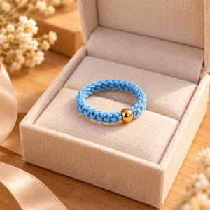 Puede incluir: Un anillo de cordón trenzado azul claro que presenta una única cuenta de metal dorado pulido. El anillo se presenta dentro de un joyero cuadrado forrado de tela beige con tapa abatible, mostrando un diseño sencillo y limpio con banda.