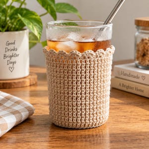 Puede incluir: Un vaso transparente lleno de una bebida helada de color ámbar con una pajita de metal, envuelto en una funda de ganchillo beige con un borde superior festoneado. La funda está tejida con un patrón de puntos denso y texturizado en hilo beige.