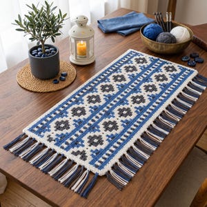 May include: A rectangular crochet table runner featuring a geometric floral pattern in white, blue, and dark gray yarn. The runner is finished with long, multi-colored fringe tassels along both shorter ends, creating a textured and decorative textile piece.