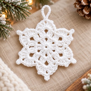 Op de afbeelding: Een wit gehaakt sneeuwvlokornament met een zespuntig sterontwerp, ingewikkelde opengewerkte steken en een klein lusje aan de bovenkant om op te hangen. Dit handgemaakte textielstuk vertoont een symmetrisch patroon met fijne garendetails.