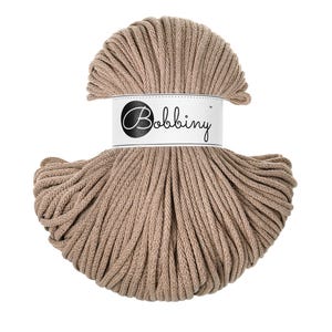 May include: A skein of light brown cord yarn featuring a braided texture. A white paper label is wrapped around the center of the yarn, displaying the brand name Bobbiny in a black script font inside a black circle on the left side.