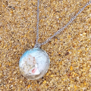 May include: A circular pendant necklace featuring a clear resin dome containing small beige and white seashells and sand particles. The pendant is attached to a silver-toned metal chain link necklace, creating a beach-inspired accessory.