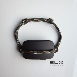 以下が含まれることがあります： カモフラージュ柄のコードブレスレットに取り付けられた黒い長方形のプレート。コードにはサイズ調整用の結び目が2つあります。画像右下のブレスレットの下には、黒い文字で「SLX DESIGNS」と印刷されています。