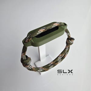May include: A camouflage patterned paracord bracelet featuring a rectangular olive green plastic housing for a fitness tracker. The cord is secured with adjustable knots on both sides. The text SLX DESIGNS is printed in black in the bottom right corner.