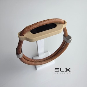 May include: A fitness tracker accessory featuring a tan, textured leather cord band held by two silver-toned metal clasps. The central housing is a matte beige plastic frame with a black rectangular screen insert. The text SLX DESIGNS is visible.