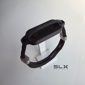 May include: A black bracelet featuring a rectangular central component with a textured surface, connected to a double-stranded black woven cord. The cord is secured by two smooth, silver-toned metal beads. The text SLX DESIGNS is visible.