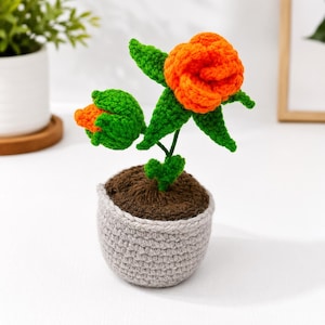 Peut inclure: Une plante en crochet en pot avec une fleur orange vif et un bouton orange plus petit, accompagnés de feuilles et de tiges vertes. La plante est fixée dans une base de terre en crochet marron foncé dans un pot gris clair texturé.