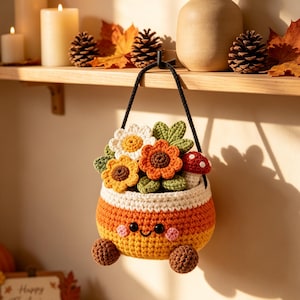 May include: A hanging crochet basket shaped like candy corn with a smiling face, pink cheeks, and two small brown feet. The basket is filled with crocheted flowers in white, yellow, and orange, green leaves, and a small red and white mushroom.