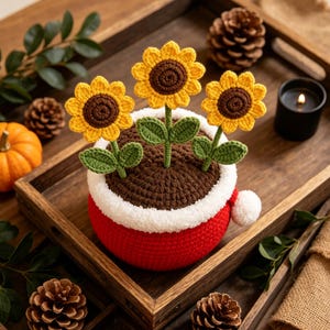 May include: A crocheted potted plant featuring three yellow sunflowers with brown centers and green leaves, set in a red pot with a white fuzzy rim and a small white pom-pom on the side, filled with brown soil-textured crochet yarn.