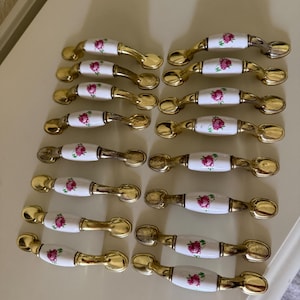 May include: A collection of fourteen drawer pulls featuring a white ceramic center with a pink rose floral pattern and gold-toned metal ends. Each handle has a curved, elongated shape with rounded mounting points on both sides of the central piece.
