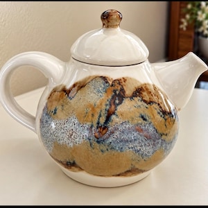 Può includere: Una teiera in ceramica con finitura lucida color bianco sporco e corpo arrotondato. Il centro presenta una fascia orizzontale di toni terra screziati, tra cui sfumature di beige, marrone e blu, con un effetto smalto testurizzato e coperchio.