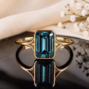 May include: A gold ring featuring a large, rectangular, emerald-cut dark blue gemstone set in a smooth bezel. The band has a split-shank design where the metal arms meet the central stone setting, creating a clean and polished appearance.