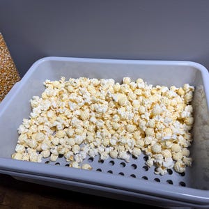 May include: A light gray rectangular plastic tray with a perforated base containing a large amount of popped white popcorn. To the left, a clear plastic container filled with yellow unpopped corn kernels is partially visible.