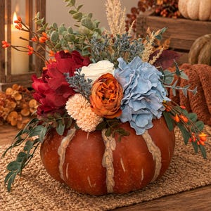 May include: A decorative orange pumpkin centrepiece filled with an arrangement of artificial flowers including deep red peonies, a white rose, a burnt orange bloom, light blue hydrangeas, small peach pom-poms, green eucalyptus, and tall dried grasses.