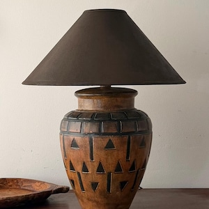 May include: A table lamp featuring a dark brown conical lampshade and a rounded base made of brown material with a carved black geometric pattern consisting of horizontal bands and triangles. The lamp has a wide neck and a tapered bottom section.