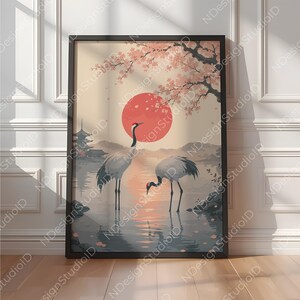 以下が含まれることがあります： 浅瀬に立つ2羽の鶴を描いた額装アートプリント。大きな赤い太陽、ピンクの花を咲かせた桜の木、そして柔らかな色調の風景を背景に、遠くにパゴダのシルエットが描かれています。