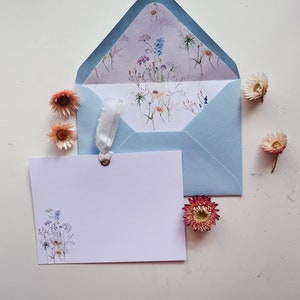 May include: A light blue envelope with a floral pattern inside. A white card with a floral design is in front of the envelope. Dried flowers are scattered around the envelope and card.