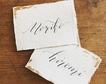 Könnte beinhalten: Zwei weiße Platzkarte mit goldenen Akzenten und schwarzer Kalligraphie. Die obere Karte sagt "Noriko" und die untere Karte sagt "Hiromi".