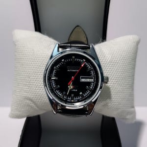 Op de afbeelding: Een Citizen automatisch polshorloge met een zwarte wijzerplaat, zilverkleurige uurmarkeringen en een rode secondewijzer. Het horloge heeft een dag-datumvenster op de drie-uurpositie, een zwarte lederen band en een klein grafisch logo boven de zes.