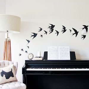 Op de afbeelding: Een zwart-witte piano met bladmuziek op de standaard. Een witte lamp met een witte kap staat links in de afbeelding. Een witte stoel met een zwart-wit kussen staat op de voorgrond. Een witte muur met zwarte silhouetten van vogels die in een zwerm vliegen is op de achtergrond.