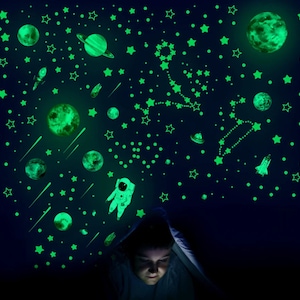 May include: A child is tucked under a blanket, looking at a glow-in-the-dark wall decal set featuring stars, planets, a rocket, a comet, and an astronaut. The decals are green and glow in the dark.