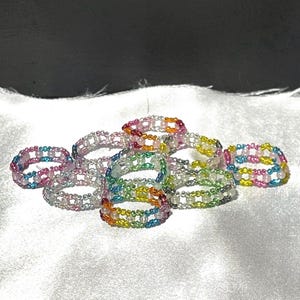 May include: A collection of handmade beaded rings featuring small, colorful glass beads arranged in a lattice pattern. The rings display various color combinations, including rainbow, pink, blue, yellow, and green, with clear beads interspersed throughout the designs.