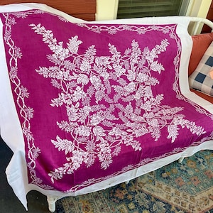 May include: A square magenta textile featuring a white botanical print of wisteria flowers and leaves. The design includes a central floral arrangement surrounded by a delicate vine border, with a solid white fabric edge framing the entire piece.