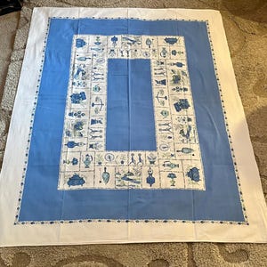 May include: A rectangular tablecloth featuring a wide white border, a solid blue inner band, and a central white panel decorated with a grid of blue and green illustrations depicting various household objects, tools, and botanical motifs.