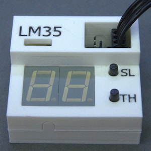 May include: A white rectangular electronic module featuring a two-digit seven-segment display, two black push buttons labeled SL and TH, and the text LM35 printed on the top left. A black wire connector is plugged into the top right section.
