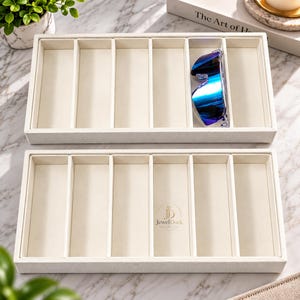 May include: Two rectangular off-white jewelry organizer trays with six vertical compartments each. The bottom tray features a circular logo with the text JewelDock. A pair of blue-tinted sunglasses rests in one compartment of the top tray.