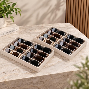 May include: Two rectangular beige storage trays, each featuring multiple compartments filled with various pairs of sunglasses. The eyewear includes frames in black, brown, and cream colors, arranged neatly within the velvet-lined organizers.