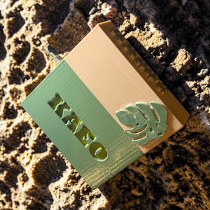May include: A square, two-toned box featuring a sage green section with the embossed text "KAYEO" and a light tan section. A small, raised, light green monstera leaf shape is attached to the tan side, and a small metal pin protrudes from the top.