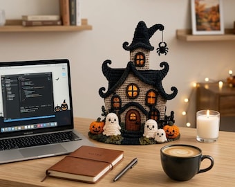 Puede incluir: Una decoración de casa encantada de Halloween tejida a ganchillo con techo negro, paredes beige y ventanas iluminadas en naranja. La estructura está decorada con tres pequeños fantasmas blancos, dos calabazas naranjas y una araña negra colgante.