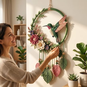 Peut inclure: Une décoration murale circulaire composée d'un colibri vert crocheté avec des accents roses, entouré de fleurs crochetées dans des tons rose, blanc et violet, ainsi que de feuilles vertes. Le cercle comporte des brins suspendus avec des feuilles.
