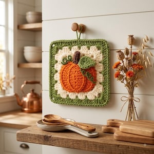 Peut inclure: Un dessous de plat carré en crochet représentant une citrouille orange avec une tige marron et des feuilles vertes, entouré d'une bordure crème et d'un cadre vert, suspendu à un crochet mural en bois dans une cuisine.