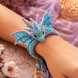 May include: A crochet dragon bracelet featuring light blue and lavender yarn, intricate wing details, and small beads. The design includes a dangling gold-tone charm with a crescent moon and a star, secured around the wrist with a wrap-around style.