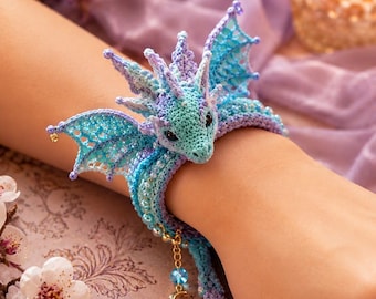 May include: A crochet dragon bracelet featuring light blue and lavender yarn, intricate wing details, and small beads. The design includes a dangling gold-tone charm with a crescent moon and a star, secured around the wrist with a wrap-around style.