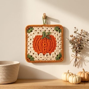 May include: A square crochet potholder featuring a central orange pumpkin motif with a green stem, surrounded by a cream-colored granny square border with green corner accents. The potholder is suspended from a small wooden hook on a wall.