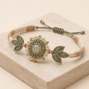 May include: A macrame bracelet featuring a central flower motif with a smooth, pale green circular stone at its core. The flower is surrounded by sage green and cream-colored knotted cord, flanked by two leaf-shaped accents and metallic beads.