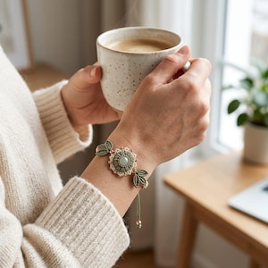 May include: A macrame bracelet featuring a central sage green circular motif with a smooth light green stone at its center, surrounded by small cream-colored beads. The design includes two leaf-shaped accents on either side of the center piece.