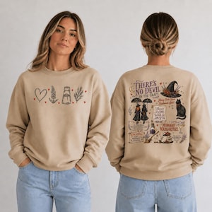 Könnte beinhalten: Ein beigefarbenes Sweatshirt mit Rundhalsausschnitt und einem Frontmotiv aus Herz, Zweig, Glas und Ast mit kleinen roten Herzen. Die Rückseite zeigt eine detaillierte Illustration von Hexen, einer Katze, einem Hut und dem Text There's No Devil In The Craft.