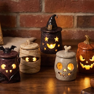 Peut inclure: Cinq pots en céramique sur le thème d'Halloween avec couvercles, chacun présentant un motif découpé éclairé de l'intérieur. La collection comprend un vampire, une momie, un pot chapeau de sorcière, un crâne et une citrouille, tous illuminés.