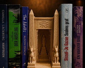 Puede incluir: Un sujetalibros de color beige con forma de entrada a un templo egipcio, con dos estatuas de chacales en la base, el símbolo del Ojo de Horus sobre el arco y una figura central en una escalinata, situado entre varios libros de tapa dura.