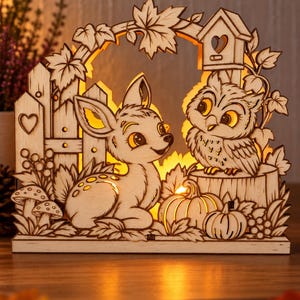 Peut inclure: Une décoration lumineuse en bois découpé au laser représentant un faon et un hibou dans une scène forestière avec des citrouilles, des champignons, une clôture et un nichoir. Le design complexe est éclairé par l'arrière, diffusant une lueur chaude.