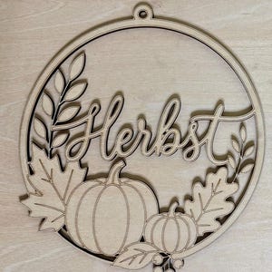 Könnte beinhalten: Ein kreisförmiger Wandbehang aus hellem Holz mit dem Wort Herbst in Schreibschrift. Das Design zeigt lasergeschnittene Details von zwei Kürbissen, Ahorn- und Eichenblättern sowie kleinen Beeren innerhalb des Rahmens.